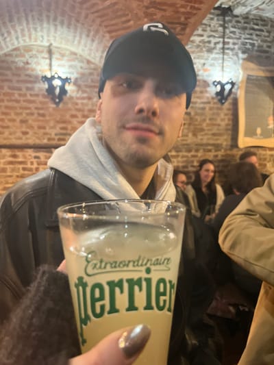A man in a black beanie and black jacket holds a glass of Perrier. His face is in focus. Behind him, people sit at tables inside a space with exposed brick walls. A decorative wall light fixture is attached to the brick. Another person is visible next to the Perrier glass, only their hand with a painted nail is visible.