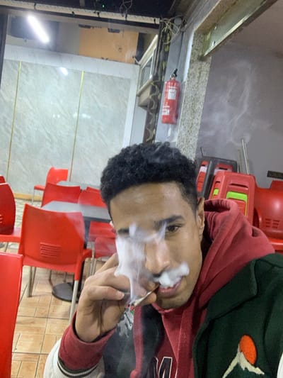 A young man with dark, curly hair exhales a cloud of smoke. He is wearing a red and green hooded sweatshirt. The background includes stacks of red chairs and a wall with horizontal gold lines. A fire extinguisher is visible on the wall.