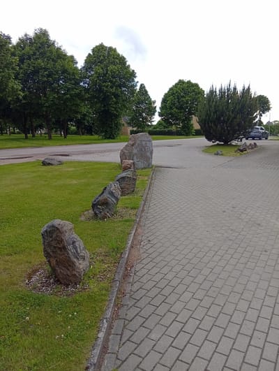 This is a ground-level, eye-level, medium shot of a paved walkway next to a grassy area with several large boulders. The walkway is made of interlocking grey rectangular pavers and curves to the right. The grassy area has scattered white clover flowers and is bordered by a curb. Several large, dark-colored boulders are placed in a line along the grassy area. One boulder, at the far left of the line, is lighter gray and has a carved relief on its surface, depicting what appears to be figures.
In the background, there are several large, mature trees with green foliage, suggesting a park or a well-landscaped area. A paved road is visible behind the grassy area and trees. Further in the background, a light-colored building with a dark roof can be seen amongst the trees, and a dark-colored SUV is parked on the right side of the road. The sky is overcast with a light grey tone.
The scene is in Micaičiai, Lithuania, indicated by the presence of the large, carved boulder, which is likely a memorial or public art piece. The overall impression is one of a public outdoor space, possibly a park entrance or a civic area, designed with natural elements like boulders and trees, alongside functional elements like the paved walkway and road. There are no people visible in the image.