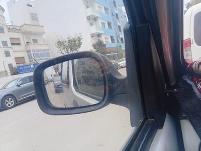 The image provides a street-level view from inside a vehicle, looking out the driver's side window and into the side mirror. The foreground features the car's black plastic door frame and a prominent side mirror.
Through the window on the left, an urban landscape unfolds under an overcast, hazy sky. Several multi-story buildings are visible, including a large white structure with balconies and a turret-like section on the far left. Below this building, a blue sign clearly displays "AL QAYM" in Latin script and "التكوين المهني" (Vocational Training) in Arabic, with "INSTITUT PRIVE DE FORMA..." partially visible, indicating a private vocational training institute. Further down the street, another building with alternating white and light blue panels is seen. Sparse, bare trees suggest a cooler season. A gray sedan, possibly an Audi, is positioned on the left side of the road, either parked or in motion.
The side mirror, framed in black plastic, reflects the scene behind the viewer's vehicle. In the reflection, a dark gray van or SUV is visible on the road, following the car. The mirror's surface appears somewhat streaked or dirty. To the far right, a portion of a white van's rear is visible outside the car window. Inside the vehicle, on the right, a colorful, patterned fabric, possibly a small rug or tapestry, is tucked into the window frame, adding a decorative element. The overall scene depicts a typical urban environment in Martil, Morocco, characterized by the architecture and visible text.