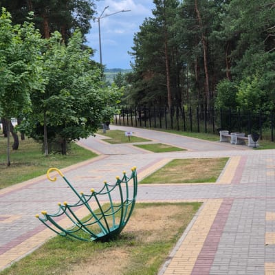The image depicts a paved park path in the city of Liski, Russia, with trees lining the right side and a fence in the background. In the foreground, a green, open umbrella-shaped metal sculpture with yellow decorative spheres is positioned on the grass. The path is constructed with interlocking concrete pavers in shades of gray, yellow, and red, forming geometric patterns. On the right side of the path, two stone benches with dark trash receptacles are visible, placed in front of the fence. A lamppost stands on the left side of the path, further back. The sky is partly cloudy.