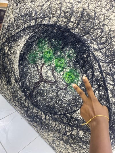 The image depicts a contemporary art piece crafted from various threads, observed in Alaguntan, Nigeria. The artwork itself is primarily composed of densely coiled black thread, forming an abstract, swirling pattern across a light-colored base material. Centrally, a tree motif is depicted with a brown thread-formed trunk and branches, and bright green coiled threads representing its foliage. A dark-skinned human hand enters the frame from the lower right, positioned over the artwork. This hand displays a thin, gold-colored bracelet on the wrist, with the index and middle fingers extended upwards in a 'V' gesture, touching or hovering just above the green foliage. A section of a white tiled floor is visible in the lower left, indicating an indoor environment where the artwork is displayed, possibly on a flat surface.