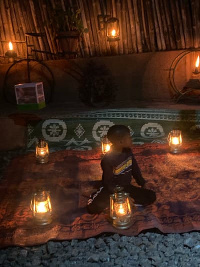 A nighttime view of an interior space in Barden Yana, Nigeria, is illuminated by multiple lanterns, casting a warm glow. The space is decorated with a patterned rug and a wooden wall. A person wearing a dark shirt with yellow and white stripes is present. The ground is covered with small rocks.