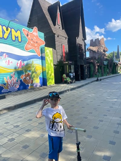 The image captures a young boy wearing a white t-shirt with a "B.Duck" cartoon and blue shorts, standing on a paved walkway. He is holding the handlebars of a scooter. To his left, a brightly colored mural depicts an underwater scene with fish and coral, and a large starfish graphic. Behind the mural, a dark, wooden, Gothic-style building stands tall, with a weathered appearance. Further back, a peculiar, large, brown, tree-like sculpture with a face is visible, adding to an unusual and theme-park-like atmosphere. A woman in a green outfit is seated in a chair near an entrance to the wooden building. The scene is set outdoors under a bright, partly cloudy sky, suggesting a sunny day. The architecture and vibrant mural suggest this location might be part of an amusement park or entertainment venue, possibly in the city of Anapa, Russia, known for its coastal attractions and family-friendly establishments.