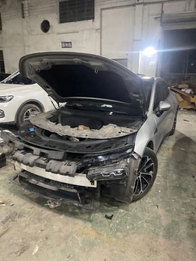 An overhead view of a damaged silver sedan with its hood open is captured in a dimly lit industrial setting, possibly a repair shop, in Panyu District, China. The front bumper and grille of the car are missing, revealing the engine compartment and various mechanical components. Debris and dirt are scattered across the concrete floor, indicating a scene of disarray. A white SUV is partially visible in the background. The lighting is stark, with a bright light source casting long shadows.