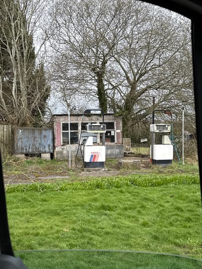The image depicts a disused petrol station in Stratford-on-Avon, United Kingdom, viewed from the interior of a vehicle. In the foreground, a vibrant green, unkempt grassy area transitions to a patch of dirt and sparse weeds. Two derelict petrol pumps stand centrally. The left pump is white with a prominent red, white, and blue "TOTAL" logo and features digital displays showing numerical values. The right pump is white, unbranded, and also exhibits numerical displays. Both pumps are equipped with hoses and appear to be non-operational. Directly behind the pumps is a dilapidated, single-story building with a corrugated metal roof and multiple window panes, some of which are broken or obscured. To the left of this structure, a rusty, weathered metal container rests on concrete blocks. The background is dominated by numerous bare deciduous trees under an overcast, light grey sky. No individuals or active vehicles are visible. The scene conveys a state of abandonment and disrepair.