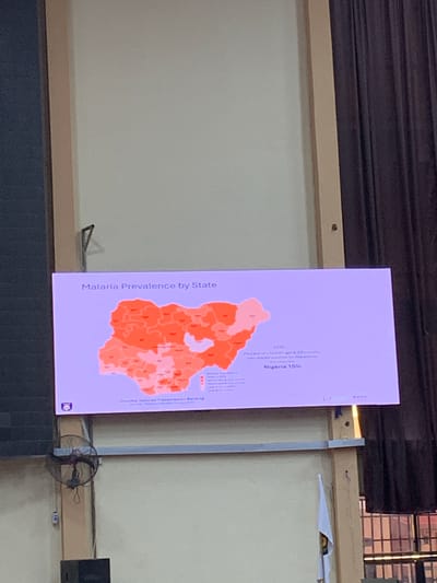 The image captures a presentation slide displayed on a screen in an indoor setting, likely in Ilorin, Nigeria. The slide, titled "Malaria Prevalence by State," features a color-coded map of Nigeria, illustrating malaria prevalence levels across its various states using shades of orange and pink.

Visible text on the slide indicates that the data pertains to the "Percent of children age 6-69 months who tested positive for malaria by microscopy." It also states "Nigeria 15%" and references the year "2025," possibly a projection or data target. A legend, though partially obscured, categorizes prevalence into ranges such as "High (> 35%)," "Moderate (15-35%)," "Low-Moderate (5-15%)," and "Low (< 5%)." Additional text mentions "Country-Tailored Transmission Banding" and likely sources the data from a "Malaria Indicator Survey 2025." The organization "PATH" is visible at the top right of the slide, along with a blue circular logo at the bottom left.

The screen is set against a light-colored wall, with a wooden beam running horizontally below it. To the right of the screen, a small electric fan with a dark grill is partially visible, suggesting a need for cooling, common in the local climate. Above the screen, dark, ribbed structures, possibly part of a ceiling or window blinds, are present. The image is taken at an angle, causing the screen to appear vertically oriented in the frame, though the text on the slide is horizontally readable. No people are visible, and the scene implies an informational session or a data review.