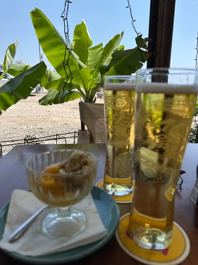 In Kissonerga Community, Cyprus, a close-up shot reveals two tall glasses of golden beer with foam heads, placed on yellow circular coasters on a dark wooden table. To the left, a clear glass dish holds a dessert, possibly ice cream with fruit, resting on a white napkin and a light blue saucer, with a silver spoon to its left. The background shows large, vibrant green banana leaves against a bright blue sky, with a gravelly outdoor area visible beyond a metal fence. String lights are draped overhead, and a blurred glimpse of buildings and vehicles can be seen in the distance. The scene suggests a relaxed outdoor dining or drinking setting.