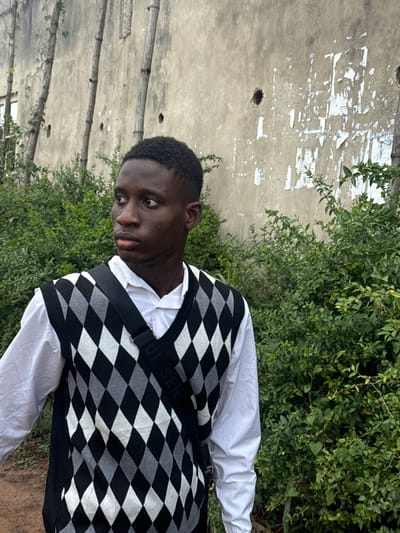 An outdoor scene in Ihiagwa, Nigeria, features a young male subject positioned slightly off-center to the right, facing and gazing towards the left. The individual has dark skin and short, dark hair. He is wearing a white long-sleeved collared shirt beneath a sleeveless V-neck argyle sweater vest patterned in black, white, and gray diamonds. A black strap from a bag, bearing white text partially visible as "UI SEN," crosses diagonally over his chest. The background consists of a weathered, light-colored concrete wall displaying several dark, circular holes and multiple remnants of torn white paper or posters. Three slender wooden poles, possibly bamboo, are vertically arranged against the wall, with one pole showing minimal vine growth. Dense green bushes and foliage surround the subject, obscuring the lower portion of the wall. A white-framed window is partially visible on the far left, behind the wall. The ground in the lower-left foreground is reddish-brown earth.