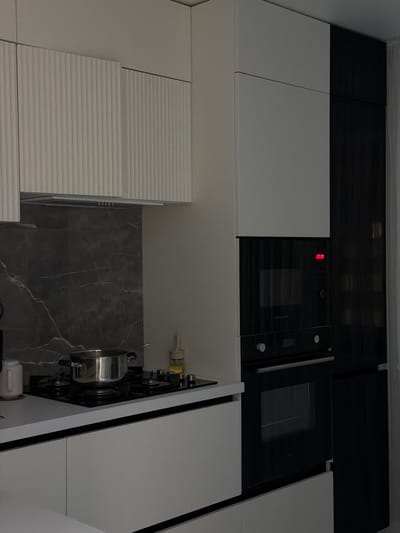 This is a wide, eye-level, interior shot of a modern kitchen, likely in Naberezhnye Chelny, Russia. The kitchen features a dual-tone design with white upper cabinets and a black appliance wall. The white cabinets have a ribbed texture and are mounted above a dark marble backsplash and a gas stovetop. A stainless steel pot sits on the stovetop, suggesting recent or ongoing cooking. Below the stovetop, white cabinets with matte finishes line the counter. To the right, a tall black unit houses a microwave and an oven, with a red digital display showing "23:00". The overall impression is one of clean lines and contemporary design, with a focus on functionality.