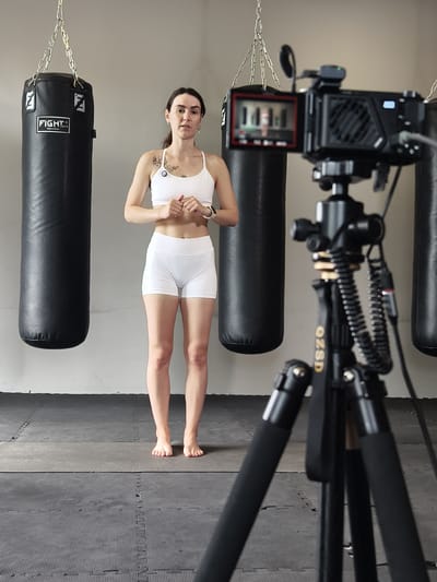 A woman in white athletic wear stands barefoot on a padded floor in a gym, facing a camera on a tripod. Two black punching bags hang behind her. The camera screen shows a blurred image of the woman. The gym has a light-colored wall and a grey floor with black matting. The image is taken from a low angle, focusing on the woman and the equipment. There is no indication of the city of Tyumen, Russia, in the image.