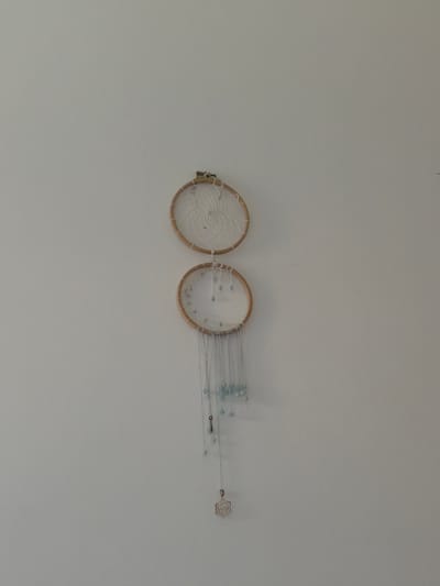 Two stacked, circular dreamcatchers hang against a plain, light-colored wall. The dreamcatchers have wooden hoops and are adorned with white string webbing and light blue beads. Dangling from the lower dreamcatcher are several thin strings with more blue beads and a silver charm at the end. The scene is indoors, likely a residential setting. There are no people or animals present, and no specific identifiable landmarks of Münster, Germany are visible.