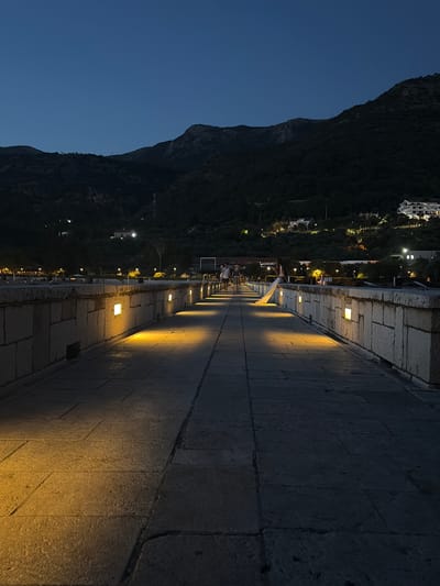 A long stone pier extends into the distance, illuminated by square lights embedded in its outer wall, casting warm glows on the paved walkway. The sky is a deep twilight blue, and dark, tree-covered mountains rise in the background, dotted with lights from buildings on their slopes. A few people are walking on the pier, silhouetted against the fading light. The scene depicts a tranquil evening in Sveti Stefan, Montenegro, possibly near the Old Town.