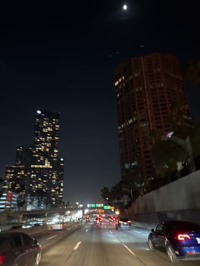 A nighttime shot of a highway in Los Angeles, California, possibly near downtown. The image features dense highway traffic with numerous vehicles and bright taillights. High-rise buildings stand tall in the background, illuminated with several lights, suggesting a bustling urban environment. The night sky is visible above the buildings, with a crescent moon hanging faintly. Overall, the scene depicts a vibrant, lively urban landscape at night with an emphasis on vehicular movement.