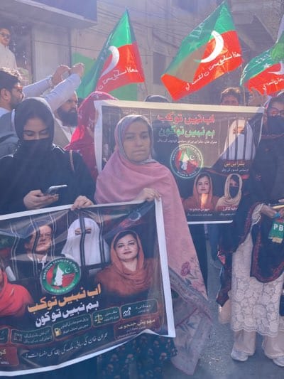 In Quetta, Pakistan, a group of people, mostly women, participate in a daytime street protest. They hold large banners featuring images of political figures and text in Urdu. Some participants carry flags with a green and red design and a crescent moon and star symbol. The environment appears to be an urban area with buildings visible in the background.