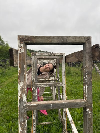 A young woman with her hair in a bun is looking into the camera and smiling. She is wearing a light-colored hooded jacket over a dark t-shirt and bright pink leggings or pants. She is positioned within a series of wooden frames, possibly old window frames, stacked one inside another. The frames are weathered and show signs of peeling paint. She is leaning forward, with one arm resting on a frame and her hand near her knee. The scene appears to be outdoors, with lush green grass and foliage visible behind the frames. The sky is overcast, suggesting a cloudy day. The orientation of the image is rotated, making the woman appear to be in a vertical position relative to the viewer. The location is identified as Rusalevka, Russia.