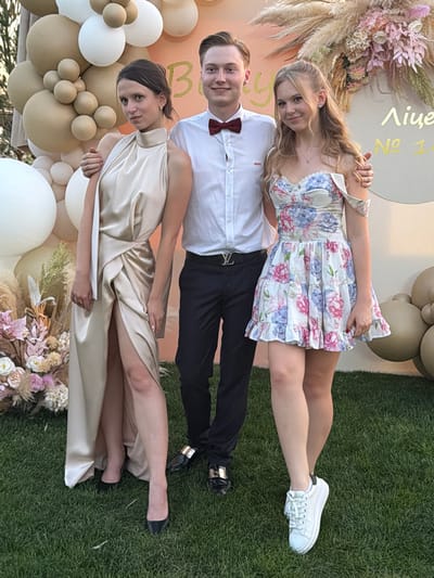 A medium shot shows three young adults posing for a photograph in an outdoor grassy area, with a beige and gold balloon arch and floral arrangement in the background. The person on the left is a woman with dark hair pulled back, wearing a long, champagne-colored satin dress with a high neckline and a thigh-high slit. She is looking directly at the camera and has one hand on her hip. In the center, a young man with light brown hair is wearing a white button-down shirt with a red bowtie and dark pants. He is smiling. On the right, a young woman with long blonde hair is wearing a floral print dress with off-the-shoulder sleeves. She is also smiling and has an arm around the man. The ground is covered in green grass, and the background suggests an event setting, likely in Khotianivka, Ukraine, given the text visible on a circular cutout behind the subjects, which appears to be in Ukrainian.