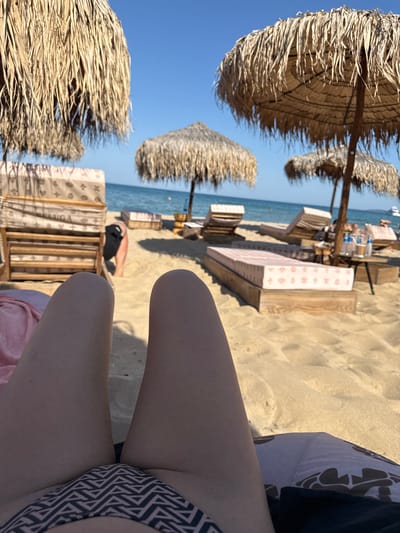 The image depicts a sunny day at a beach in Sozopol, Bulgaria. The scene is viewed from a low angle, focusing on the legs of an individual sitting on the sand, wearing a patterned bikini bottom and a dark top. In the background, several thatched beach umbrellas provide shade over rows of loungers, some with patterned covers. Beyond the loungers, the calm blue sea stretches out under a clear blue sky. The sand appears dry and light-colored, with visible footprints and indentations. A person in a black shirt is visible lying down further in the background. The time of day is likely midday or early afternoon, given the bright sunlight casting shadows. The overall mood is relaxed and vacation-like. The text "LUGA" is visible on one of the lounger covers.
