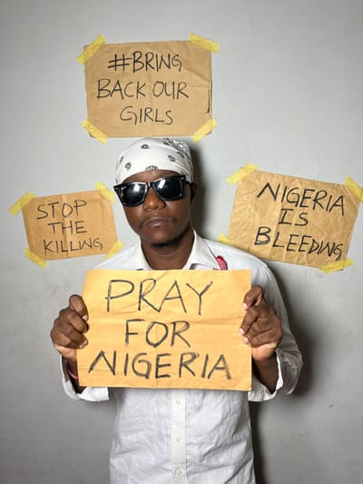 A person lies on a gray surface surrounded by four handwritten signs. The signs, attached with yellow tape, say: "Nigeria is Bleeding," "#Bring Back Our Girls," "Stop The Killing," and "Pray For Nigeria" (which the person holds). The person wears a white shirt, a bandana, and sunglasses. The signs are on brown paper.