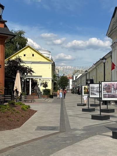 The image depicts a sunny day on a pedestrian street in Putyatino (Semivragovskaya S/A), Russia. The street is lined with buildings, one a bright yellow structure on the left and a longer, pale yellow building with white trim on the right. Decorative garlands of what appear to be musical notes are strung across the street overhead.
In the foreground, on the left side, is a statue of a person seated on a bench, surrounded by landscaping. Adjacent to this is a covered outdoor seating area with tables and chairs, and a large, light-colored umbrella.
The pavement is made of large, gray rectangular stones. A black metal grate runs along the edge of the sidewalk on the right side.
Several large framed artworks are displayed on black stands along the right side of the street. These artworks appear to be paintings of landscapes and architecture.
A few people are walking along the street. On the right side, a person wearing a white skirt and top is looking at the artwork. Further down the street, a group of three people, two adults and a child, are walking away from the camera. On the far left, two figures are visible in the background.
The sky is blue with scattered white clouds. Trees with dark green foliage are visible in the background, particularly behind the yellow building.