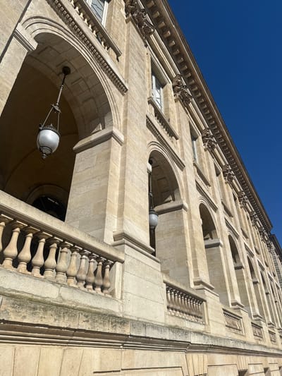 A low-angle, upward-looking shot depicts a section of the Palais Rohan, Bordeaux City Hall, in Bordeaux, France. The building is constructed from light-colored ashlar masonry, presumed to be limestone, showing signs of weathering and discoloration at the base of its structures. A stone balustrade with multiple balusters is visible in the foreground. Behind this, a series of large, rounded arches forms a loggia, supported by robust rectangular piers. Within two of these arches, spherical, white-globbed lanterns are suspended by decorative dark metal fixtures. The upper facade features rectangular windows flanked by classical architectural elements, including ornate corbels or capitals, beneath a pronounced cornice. The upper right quadrant of the image is occupied by a clear, deep blue sky. The scene is devoid of people, actions, or transient objects, illuminated by direct sunlight that casts distinct shadows under the arches.