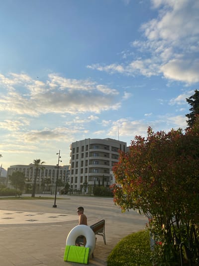 This media file captures an outdoor scene in what appears to be a resort or residential complex, likely in Sirius, Russia. The image is oriented vertically, with the sky taking up a significant portion of the frame. Wispy white clouds are scattered across a bright blue sky, suggesting fair weather, possibly late afternoon or early evening given the warm light.
In the foreground, a paved area with a decorative pattern is visible. A person, seemingly male and shirtless, is standing next to a white inflatable ring and a bright green object, possibly a mat or towel. The person is looking downwards, their posture relaxed. To the right of the person, a bench is partially visible, and beyond that, manicured green grass.
In the mid-ground and background, several modern buildings with multiple stories and numerous windows are prominent. These structures have a contemporary architectural style. Trees with reddish-tinged leaves are also present, adding a touch of color to the scene, as are palm trees, indicating a potentially mild climate. The overall impression is one of leisure and relaxation in a well-maintained urban or resort environment. There is no visible text in the image.