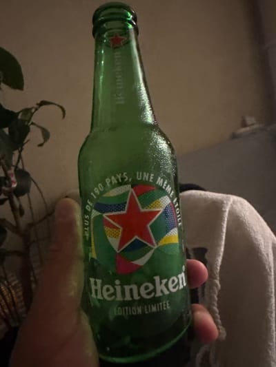 A hand holds a green glass bottle of Heineken beer, which is covered in condensation. The label on the bottle features a red star and text that reads "PLUS DE 190 PAYS, UNE MÊME ÉTOILE" and "EDITION LIMITÉE". The background is out of focus and appears to be an indoor setting with dim lighting. There is a plant on the left side of the image and a textured fabric on the right. No specific location in the city of Nice, France can be identified from the image.