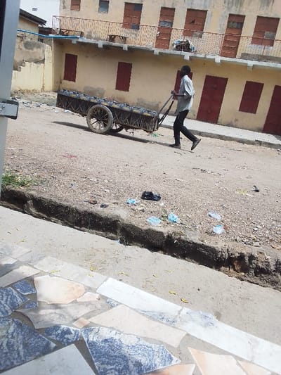 The scene depicts an outdoor urban environment in Sabon Gari West, Nigeria. A dark-skinned adult male, wearing a light-colored short-sleeved shirt, dark trousers, and dark shoes, walks from right to left, actively pushing a two-wheeled, unmotorized metal cart. The cart is heavily loaded with multiple dark-colored rectangular containers, appearing to be jerry cans, arranged in rows.

The ground is an unpaved, light brown dirt road. A distinct, shallow dirt drainage ditch or curb runs parallel to the road on the right side. Scattered refuse, including light blue and white plastic bags and a black plastic bag, is visible on the ground and within the ditch.

In the background, a two-story building with a light beige or yellow facade occupies the mid-ground. Its upper floor features a metal balcony railing and five prominent, rectangular windows or doors with dark reddish-brown panels. The lower floor also displays several similar reddish-brown panelled openings. A blue pipe or conduit is affixed to the building's exterior on the left. Partial views of additional structures are visible further left.

The immediate foreground features a tiled surface, composed of irregularly shaped, multi-colored (grey, white, beige, brown) stone or ceramic pieces, suggesting a pavement or entryway. Sparse green vegetation is visible along the left edge of the dirt road. The scene is brightly lit by sunlight, casting visible shadows.