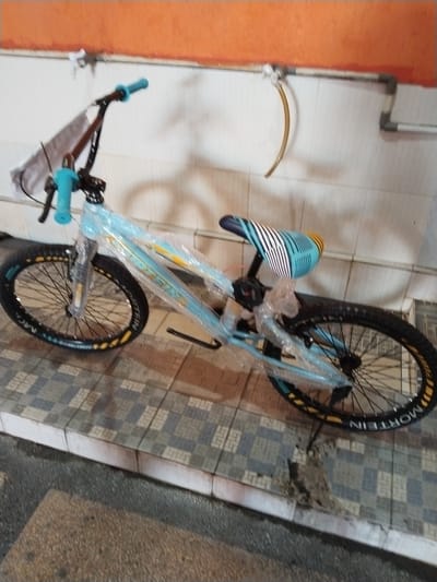 A light blue BMX bicycle, partially wrapped in protective plastic, is positioned leaning against a tiled wall with an orange stripe at the top. The bicycle has black handlebars with blue grips, a blue seat with a striped pattern, and black wheels with yellow lettering on the rims. The bike appears to be in a storage or workshop area, indicated by the tiled floor and exposed piping on the wall. The overall impression is that of a new or recently purchased item. The setting is indoors, possibly a shop or garage, and the location is identified as Pekanbaru, Indonesia.