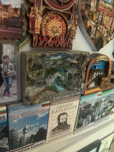 The image displays a detailed close-up of a collection of souvenir magnets and miniature book-shaped items arranged on a plain, light-colored vertical surface. In the upper section, a large, ornate multi-layered magnet resembling a clock or gear features relief figures and a central design with red and yellow patterns. The word "PRAHA" (Prague) is prominently displayed in red, three-dimensional letters at its base. To its right, a glossy, relief magnet shows a stone arched bridge over water with numerous buildings on a hillside, indicative of a European city. A portion of another blurred magnet with building imagery is visible in the top left. The middle section features: 1. On the far left, a rectangular magnet or frame holding a color photograph of a young boy, approximately 8-12 years old, with short dark hair, wearing a light grey hooded jacket, blue jeans, and red sneakers. He stands with crossed legs, holding a pair of black binoculars, against a blurred background. 2. Centered, a rectangular relief-sculpted magnet depicts a natural scene of a cave entrance with a waterfall, rocky cliffs, and green foliage. Below the relief, the Cyrillic text "ДЕВЕТАШКА ПЕЩЕРА" (Devetashka Cave) is visible. 3. On the right, a distinctive wagon-shaped magnet with an arched top banner reading "BULGARIA" and smaller, partially legible Cyrillic text including "музей" (museum) and "Тодор Каблешков" (Todor Kableshkov). The main body of the magnet illustrates a historic building and a large wooden wheel. Below this, the Cyrillic text "КОПРИВЩИЦА" (Koprivshtitsa) is visible. The lower section consists of several small, book-shaped items, likely magnets, standing upright. Each features a design with stripes in the colors of the Bulgarian flag (white, green, red). 1. The second item from the left is titled "ПАМПОРОВО" (Pamporovo) below "ИЗДАТЕЛСТВО БЪЛГАРИ" (Bulgarian Publishing House). Its cover illustrates a tall TV tower amidst snow-covered pine trees under a cloudy blue sky. Below, "КОЛЕКЦИЯ КНИЖКИ" (Booklet Collection) is written. 2. The third item is titled "ЛЮБЕН КАРАВЕЛОВ" (Lyuben Karavelov) below "ИЗДАТЕЛСТВО БЪЛГАРИ". Its cover displays a black and white portrait of a bearded man, Lyuben Karavelov. Smaller text reads "ИЗБРАНИ ПРОИЗВЕДЕНИЯ" (Selected Works) and "МАГНИТНА БИБЛИОТЕКА" (Magnetic Library). 3. Parts of additional book-shaped items are visible on the far left and right, and between the Pamporovo and Lyuben Karavelov items, showing figures in historical attire. The items are static, displayed indoors, possibly on a magnetic board or refrigerator. No information regarding the location "Pokrovnik,