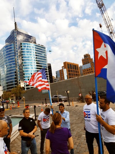 A group of people stand outside, holding flags of the United States and Cuba. They wear shirts with slogans referencing freedom for Cuba. Buildings rise in the background, including a modern glass skyscraper and brick structures. A construction crane is visible. The atmosphere is one of protest or demonstration, with the flags and clothing expressing political sentiments.