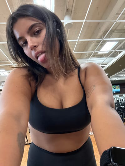 A young adult female is depicted in a selfie, taken in a fitness center located in Plainsboro Township, United States. She has dark hair, a medium skin tone, and is wearing a black sports bra and black athletic bottoms. Her left arm, extended towards the camera, shows a minimalist line-art tattoo on the upper bicep and a dark smartwatch on the wrist. She is looking directly into the camera with her tongue slightly protruded. The background features a grid-patterned dropped ceiling with integrated artificial lighting, a light-colored wooden floor, and blurred exercise equipment, including a distant television screen, consistent with a gym environment.