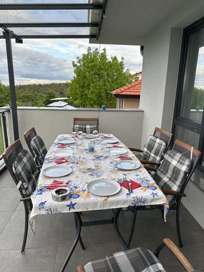 The image depicts a set outdoor dining table on a balcony in Brestnik, Bulgaria. The table is covered with a white tablecloth with a sea-themed design including coral, starfish, and shells. Six place settings are arranged, each consisting of a white plate, silverware, a red napkin, and several glasses. An ashtray sits near one of the place settings. Six chairs with gray and white plaid cushions surround the table. The balcony is enclosed by a metal and glass structure, offering a view of a tree, a house with a red-tiled roof, and a forested area in the distance under a cloudy sky.