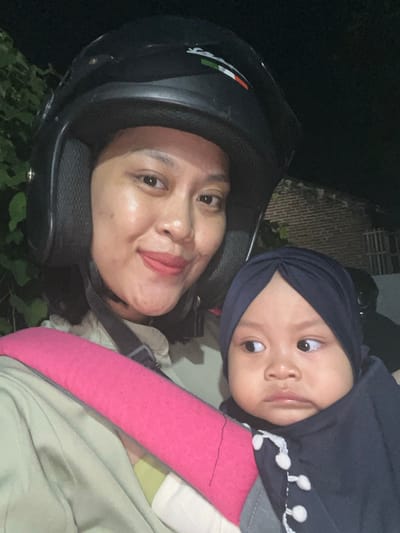The image portrays an adult female and an infant in close proximity, situated in Kecamatan Pati, Indonesia, during nighttime. The adult female, positioned on the left and center, wears a black motorcycle helmet adorned with a white, green, and red stripe logo. She has reddish lipstick and a slight smile, looking directly at the viewer. She is carrying an infant, visible in the lower right, secured in a baby carrier featuring a bright pink padded shoulder strap. The infant wears a dark blue turban-style head covering and a dark blue garment with white decorative accents on the front. The infant's gaze is directed towards the right, away from the viewer. The background is dimly lit, revealing dark green foliage on the left and a reddish-brown brick wall structure with a visible opening on the right.