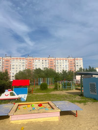 This is a vertical, eye-level, outdoor photograph taken on a sunny day with a partly cloudy sky. The scene is a children's playground with a large, apartment building in the background. The building appears to be a typical Soviet-era multi-story residential building, with multiple windows and balconies. Trees and green grass are visible in front of the building, suggesting a summer or early autumn season.
In the foreground, there is a sandbox filled with sand and various colorful toys, including a red bucket, blue toys, yellow toys, and green toys. Adjacent to the sandbox is a part of a playground structure, painted in blue and red with white accents, designed to resemble a vehicle, possibly a boat or a truck, with a steering wheel and wheels. Another playground structure, made of metal tubes in red, blue, and yellow, is visible behind the sandbox.
To the right of the sandbox, a blue structure with a checkerboard pattern on a square panel is visible. The panel has some visible text in the form of a grid, with letters (A, B, C, D, E, F, G, H) along the top and numbers (1, 2, 3, 4, 5, 6, 7, 8) along the side, resembling a chessboard coordinate system. There are also some power lines visible against the sky. The overall impression is a typical urban playground in Russia, likely in the city of Magnitogorsk, given the context. No people are visible in the photograph. The lighting suggests it is daytime, likely late morning or early afternoon.