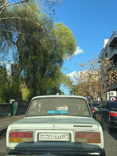 The image depicts a daytime street scene from the perspective of a vehicle, in Damascus, Syria, specifically within the Al-Muhajerin Municipality. The primary subject is the rear of a light-colored (possibly white) older model sedan, observed from a slightly elevated position.
The sedan's rear windshield features a rectangular blue and white sticker displaying the word "ARGENTINA" in black capital letters, positioned above a smaller graphic with two horizontal light blue stripes bordering a white stripe. A reflective object, possibly an air freshener or ornament, is visible behind the sticker. The vehicle's rear panel includes a centrally mounted Syrian license plate, which reads "دمشق S 888726" in Arabic script and "٨٨٨٧٢٦" in Eastern Arabic numerals. The taillight assemblies, consisting of red and amber sections, are present on both sides.
To the left of the light-colored sedan, a green, low-height fence or barrier runs parallel to the street, backed by dense green foliage. A large tree with numerous light green, drooping leaves dominates the left side of the frame. To the right of the sedan, a queue of vehicles is visible, including a black sedan directly behind the white car. Further along the street on the right, a multi-story building with balconies is present, alongside a tree with sparse, brown leaves. A "P" sign, likely indicating parking, is visible on the right sidewalk.
The sky is clear blue with scattered white cumulus clouds. No individuals are explicitly visible in the frame. The overall environment suggests an urban or suburban setting with a mix of natural elements and buildings, indicating a normal day with stationary or slow-moving traffic.