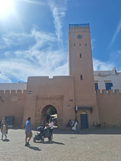 In Essaouira, Morocco, a tan-colored clock tower stands tall against a bright blue sky with scattered white clouds. The tower has a decorative blue railing at the top and a circular clock face with Roman numerals. Below the clock, there are several small, rectangular windows. The tower is attached to a fortified wall with crenellations, made of the same tan material. In the foreground, a cobblestone plaza is visible. Several people are scattered throughout the plaza. To the left, a woman in a light-colored jacket and pants is walking away from the camera. In the center, a man in a blue shirt and shorts walks towards the archway. Next to him is a double stroller with a person pushing it. To the right of the archway, a woman in a white shirt and dark pants is standing and looking at her phone. Further to the right, a bicycle is parked near a building with blue shutters.