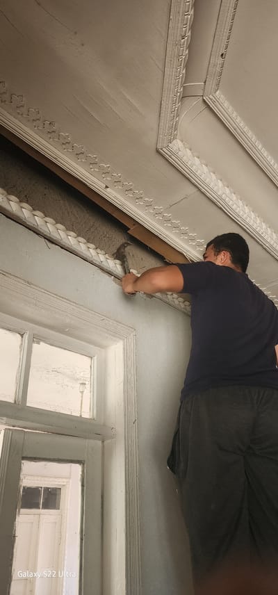 The image depicts an interior scene within a building, likely a residential or older commercial structure. A young man, wearing a dark t-shirt and dark shorts, is actively engaged in demolition work. He is holding a hammer and appears to be prying away a piece of molding or trim from a ceiling or upper wall. The area he is working on is ornate, with decorative plasterwork visible along the ceiling edge and a patterned trim running horizontally.
Adjacent to the demolition work is a window frame, also adorned with decorative trim. The window itself has multiple panes, and through it, another interior space with closed doors and what appears to be a light fixture is reflected. The walls and ceiling are painted in a light, neutral color, showing signs of age with some texture and potential imperfections. The overall impression is one of renovation or repair in an older building. The presence of the "Galaxy S22 Ultra" watermark at the bottom left indicates the image was captured with a Samsung smartphone.
