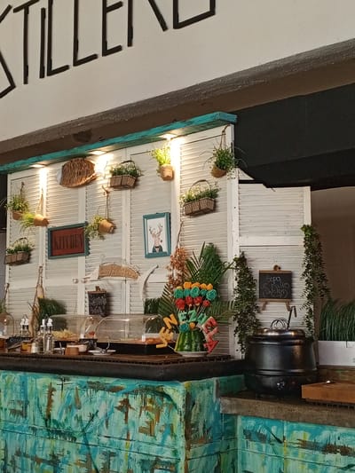 The image shows a buffet setup at what appears to be a restaurant or eatery. The background features a white wall with louvred doors, adorned with various decorative items including woven baskets, potted plants, framed pictures, and a decorative fish made of straw. The text "KITCHEN" is visible on a framed sign.
In the foreground, a buffet counter displays food items under clear covers. Bottles of condiments and other serving ware are also present. To the right, a black soup tureen is placed on the counter, suggesting a hot soup offering. A small chalkboard sign on an easel next to the soup tureen reads "Sopa de Pollo (Chicken Soup)". A colorful arrangement of what looks like decorative flowers or sweets is placed on the left side of the buffet. The counter itself is painted with a vibrant, abstract pattern in shades of teal and brown.
The lighting suggests it could be daytime or evening, with artificial lights illuminating the buffet area. The overall atmosphere seems casual and inviting, typical of a dining establishment aiming for a rustic or eclectic decor. The location context of Porlamar, Venezuela, does not add specific visual cues to the scene itself but places it geographically. There are no people visible in the image.