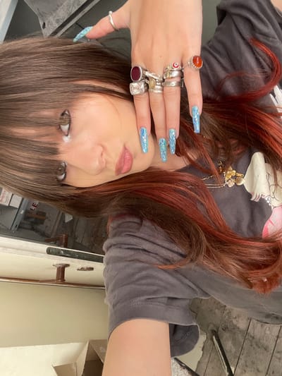 This is a close-up, upside-down portrait of a young woman with long, reddish-brown hair. She is wearing a dark gray t-shirt. Her hands are adorned with multiple silver rings, some featuring stones, and her fingernails are painted with a holographic blue design. One ring on her left hand has a circular orange stone, and another has a red stone. Her face is tilted, with her eyes looking towards the camera. She has light-colored eyes and is wearing makeup, including mascara and lip gloss. The background appears to be an interior space with wooden flooring and a wall with a door frame. A cardboard box is visible in the background, and some industrial-looking elements are present, suggesting a workshop or unfinished space. The lighting suggests it might be daytime, but the indoor setting makes it difficult to determine the time of day definitively. No specific event or activity is clearly depicted, other than the pose for a selfie. The location context of Raanana, Israel, does not provide any immediately discernible visual cues within the image itself.