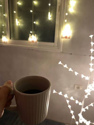 This image depicts an indoor scene at night in Nuseirat, Palestinian Territory. A light-skinned human hand, positioned in the lower-left foreground, holds a white ceramic mug with vertical ridges, filled with a dark liquid. In the background, a white-framed window is visible, adorned with illuminated string lights featuring multiple mosque-shaped ornaments and individual small bulbs. To the right of the frame, a decorative, branched light fixture, resembling a small tree, glows with numerous small, star-shaped or floral-shaped white lights. The overall environment is dimly lit by the warm, yellow-white glow of the decorative lights, illuminating light-colored walls.
