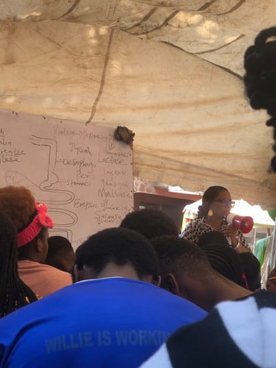 The scene depicts an outdoor educational or informational gathering under a makeshift, light-colored canvas shelter in Akpaka Forest Reser, Nigeria. A woman, positioned on the right, wears glasses, large hoop earrings, and a patterned top, actively speaking into a red megaphone. She addresses an unseen audience, whose backs and heads are visible in the foreground and midground, indicating they are facing her and a whiteboard.
The whiteboard, located on the left, displays handwritten biological diagrams and text related to the human digestive system, including terms such as "Protein -> Peptones," "Glycerol," "Oesophagus," "Stomach," "Lactase," "Maltase," "Sucrose," "Glucose," and "Erepsin." A small, dark, undefined object is attached to the top right corner of the whiteboard. Audience members include individuals with varied hairstyles, one wearing a pink headband, and another prominently in the foreground wearing a blue shirt with the white text "WILLIE IS WORKING" printed on the back. Daylight illuminates the scene, with some distant green foliage and structural elements visible beyond the shelter.