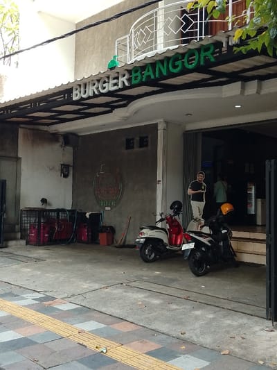 The image shows the exterior of a restaurant named "Burger Bangore" located in Pati, Indonesia (latitude: -6.753, longitude: 111.04). The restaurant has a sign above the entrance with the name in green letters. The sign is mounted on a metal frame and is partially covered by a white awning. The awning is supported by black metal beams. The restaurant has a white exterior with a gray concrete wall behind the sign. The wall has a large sign with the word "Burger" in red letters. The sign is framed in black and gold. There are two motorcycles parked in front of the restaurant.  A man in a white shirt and jeans is standing next to the motorcycles. Another man wearing a red shirt and dark pants is standing to the left of the first man. Both men appear to be waiting for something. There are several red gas tanks and a green broom leaning against the wall.  A small metal gate to the left of the entrance is partially open. The ground in front of the restaurant is covered in gray concrete.  The sidewalk has yellow painted lines.