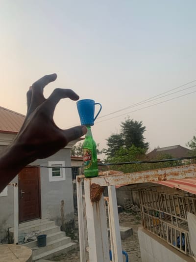 The image, correctly oriented, depicts a dark-skinned hand carefully balancing a light blue plastic cup upside down on top of a green glass bottle. The bottle, which has the word "TROPHY" prominently displayed on its label, is standing upright on a rusty, white metal railing or gate structure.
The scene is set outdoors, likely in a residential area of Kurudu, Nigeria, during what appears to be late afternoon or early evening due to the hazy, pale sky and soft light. Overhead, utility wires stretch across the frame. In the background, green trees are visible, along with residential buildings. One building is a light grey structure featuring a brown door and a white-framed window. Concrete steps lead down towards this door from the level of the railing. The rusty nature of the metal structure adds a detail to the setting. The focus is on the delicate act of balancing the cup and bottle.