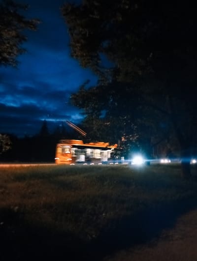 An out-of-focus, nighttime photograph taken from a low angle shows a streetcar, identified as being in Poltava, Ukraine, moving from left to right across a grassy median. The streetcar is predominantly orange with yellow accents and has two poles extending to overhead wires. The background features a dark, cloudy sky with a deep blue hue. Trees frame the top of the image, with one large tree on the right casting shadows. Several cars are visible in the distance on the right side of the frame, with their headlights creating bright, blurred streaks of light. The foreground consists of grass, also appearing somewhat blurred due to the motion of the streetcar and the low light conditions. The overall impression is one of movement and the ambiance of a city at night.