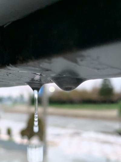 A close-up shot features a dark, likely metallic, object with a protruding round feature. Water is seen emerging from the object, forming a thin, horizontal stream. The background is blurred, showing glimpses of white buildings, green trees, and a light-colored pathway, suggesting an outdoor setting with snow or light-colored surfaces. The overall scene appears to be taken during a cold or wintry day.