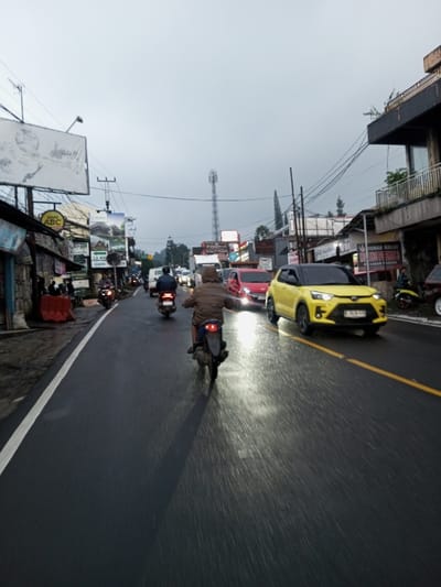 The image depicts an overcast street scene in Dawathang_Dorjibi_ Kashingtsawa, Bhutan, characterized by a wet asphalt road indicating recent precipitation. The perspective is from behind a moving motorcycle, focusing on traffic flow. In the immediate foreground, a person wearing a brown puffer jacket rides a dark-colored motorcycle away from the viewer, with the motorcycle's red tail light visible. Their arms are extended, holding the handlebars. Ahead, two more motorcycles with riders are visible. On the right side of the two-lane road, a bright yellow SUV with a black roof (likely a Toyota Raize or Daihatsu Rocky) is moving towards the viewer, its headlights illuminated and reflecting on the wet surface. Behind it, a red compact car also approaches with illuminated headlights. Further down the road, additional vehicles, including a white van, are caught in what appears to be moderate traffic congestion.

The road features a solid white line marking the left edge and a double yellow line (one solid, one broken) indicating the center division. Both sides of the street are lined with commercial buildings, many displaying various signs. On the left, signs include "KOPI ABC" and one indicating "1.2 KM". Orange and white traffic barriers are present on the sidewalk. On the right, a multi-story building features balconies with sparse greenery and signs, one partially legible as "RUMAH MAKAN" (restaurant). Overhead, a network of utility wires and poles is visible. In the background, a tall cellular tower stands prominently against the grey sky, with distant hills or mountains partly obscured by haze or low clouds. Several evergreen trees are scattered throughout the background.