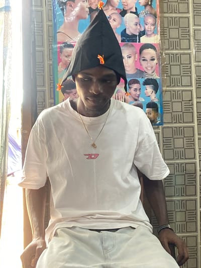 A young Black male sits facing the camera, looking down. He wears a black, pointed hat adorned with small orange flowers. He has on a white t-shirt with a pink diesel logo on the chest and a gold chain with a small pendant. Light streams in from the left, illuminating his arm and the right side of his face. Behind him, a wall is covered with a collage of images of women with short hairstyles. The floor is tiled with a patterned design. The scene appears to be indoors, possibly a salon or a personal space, in Kuson, Nigeria.