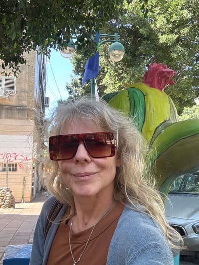This selfie captures an adult with long, wavy blonde/grey hair, wearing large brown tortoiseshell sunglasses, a brown top, and a blue cardigan. The person displays a calm, slightly smiling expression. A silver chain is visible around their neck.

Behind and above the person is a prominent, large sculpture resembling a stylized cactus or fruit, painted in vibrant lime green and darker green with black outlines, and topped with a red, rose-like element.

The setting is an urban outdoor environment under bright daylight, suggesting a sunny day. Dense green tree foliage frames the left side and upper background. A turquoise street lamp is partially visible through the trees on the left. In the background, various buildings with light-colored facades (beige, grey, and white) are seen, some with windows and an air conditioning unit. A partial view of a silver car is in the upper right. On a wall in the lower background, red graffiti or scrawled text is visible. A blue fabric object, possibly an awning or banner, hangs in the mid-left. The ground appears to be paved.

The image is a selfie, showing the person posing with the sculpture in what appears to be a public street or square in Ramat Gan, Israel. The image itself is rotated 90 degrees counter-clockwise.
