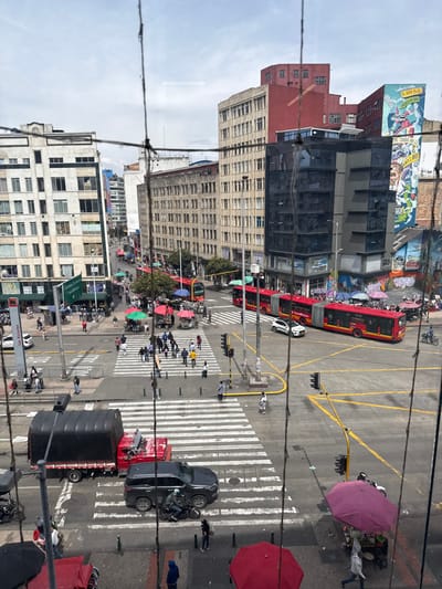 This image depicts a high-angle view of a busy urban intersection in Bogota, Colombia, specifically at the junction of Carrera 7 and Calle 20, identifiable by a visible street sign indicating "Carrera 7" and "Calle 20 Estación Las Nieves." The sky is overcast.
The intersection features marked crosswalks. Traffic includes multiple red articulated TransMilenio buses, a dark grey SUV, a red flatbed truck with a black tarp-covered load, a white SUV, and several motorcycles. Vehicles are observed stopped or navigating the intersection.
Numerous pedestrians are visible on sidewalks and actively crossing the streets within the designated crosswalks. Several street vendors are present, operating under colorful umbrellas (red, pink, green, blue) on the sidewalks, with some associated with small pushcarts.
The surrounding environment consists of multi-story buildings displaying varied architectural styles, ranging from older, light-colored structures to modern buildings with dark reflective facades. A prominent large mural featuring colorful, cartoon-like characters and text is visible on the side of a building on the right side of the image. Traffic lights are operational at the intersection, and overhead utility lines crisscross the scene, with several prominent wires appearing in the immediate foreground, partially obscuring the view.